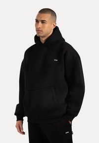 Mann mit kurzen Haaren, der einen schwarzen Hoodie und passende schwarze Jogginghosen trägt, steht mit entspannten Händen vor einem schlichten, hellen Hintergrund.