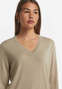 Beige V-Ausschnitt-Pullover mit langen Ärmeln, glatter Textur und einem subtil gerippten Kragen, getragen mit goldenen Creolen.