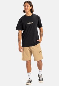 Camiseta de algodón negra con logo blanco, pantalones cortos de cargo caqui, calcetas blancas con rayas grises y zapatillas negras. Estilo de moda urbana casual.
