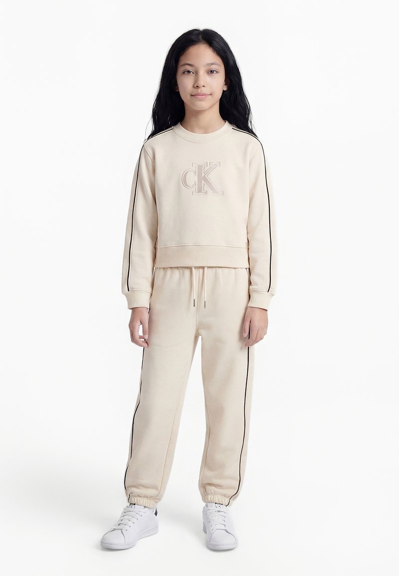 Fiatal személy hosszú sötét hajjal, bézs színű Calvin Klein pulóvert és hozzáillő fekete csíkos melegítőt, valamint fehér tornacipőt viselve, állva.
