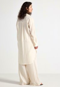 Camicia lunga di colore crema, ampia e informale, con un tessuto testurizzato, caratterizzata da un orlo dritto e polsini con bottoni, abbinata a pantaloni larghi coordinati.