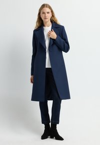 Cappotto lungo blu navy con una texture trapuntata, ampi revers, chiusura con un bottone singolo e tasche laterali, abbinato a jeans scuri e stivali neri.