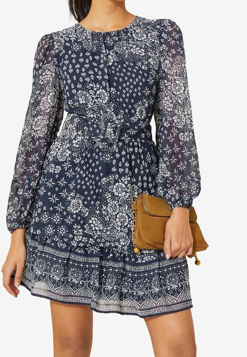 Navy blue floral print dress with long sheer sleeves and belted waist, paired with a small brown suede clutch bag.