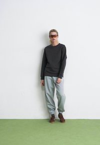 Persona che indossa un felpa scura, pantaloni della tuta grigio chiaro, scarpe da ginnastica marroni e grandi occhiali da sole, in piedi contro una parete bianca su un pavimento verde.