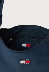 Geantă Tommy Jeans de culoare bleumarin, cu inscripții vizibile ale logo-ului pe față și în interior, arătând un buzunar cu fermoar deschis în interiorul genții.