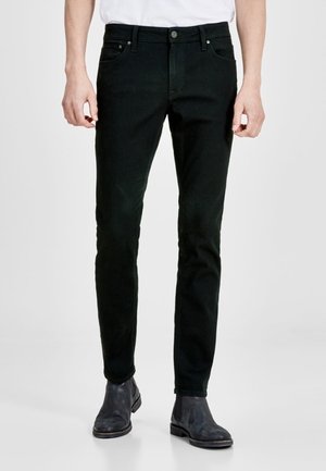 Mężczyzna w czarnych, dopasowanych jeansach i ciemnych skórzanych botkach, stojący na tle jednolitego jasnego tła.