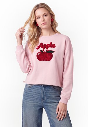 Ung kvinde med langt blondt hår iført en pink sweatshirt med rødt æblemotiv og blå jeans mod en hvid baggrund.