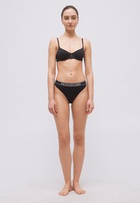 Γυναίκα όρθια, ξυπόλυτη, με μαύρο σουτιέν και μαύρο εσώρουχο Polo Ralph Lauren, κοιτάζει μπροστά, μπροστά σε απλό φόντο.