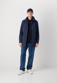 Giacca impermeabile blu navy con colletto alto, bottoni frontali e texture liscia, abbinata a jeans blu larghi e sneakers bianche.