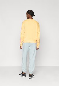 Κίτρινο φούτερ με ροζ λεπτομέρειες, χαλαρή εφαρμογή, συνδυασμένο με ανοιχτό μπλέ παντελόνι φόρμας. Αθλητικά παπούτσια μαύρα με λευκές και τρεις ρίγες.