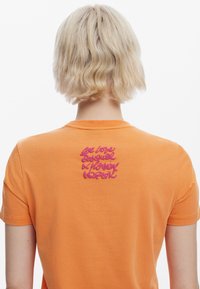 T-shirt arancione a maniche corte con testo ricamato rosa sul retro, con scollatura rotonda e texture in cotone morbida.