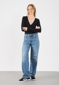 Camisola preta com riscas, de mangas longas e decote em V profundo, combinada com calças largas de ganga azul claro e ténis pretos com riscas brancas.