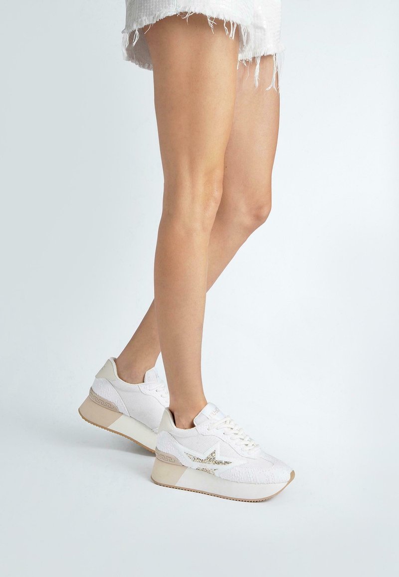 Scarpe da ginnastica bianche con una parte superiore in tessuto texturizzato, caratterizzate da un dettaglio a forma di stella scintillante e un accento beige su una suola a zeppa.