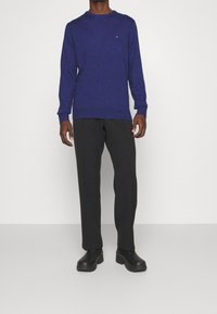 Maglione blu da uomo con maniche lunghe e colletto a costine, abbinato a pantaloni neri e scarpe nere, su uno sfondo semplice.