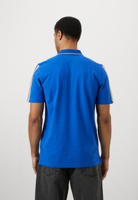 Μπλε πόλο μπλούζα με υφή, κοντά μανίκια και κλασικό γιακά. Διαθέτει κρεμ λεπτομέρειες κατά μήκος των ώμων. Εμφανίζεται πίσω.