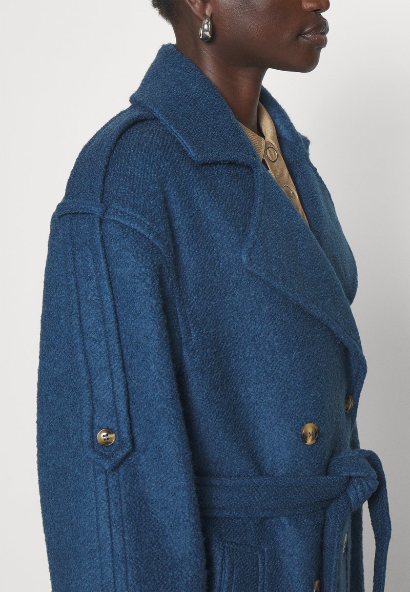 Niebieski płaszcz z mieszanki wełny, z wykładanym kołnierzem, podwójnym zapięciem, złotymi guzikami oraz materiałowym paskiem związanym w talii.