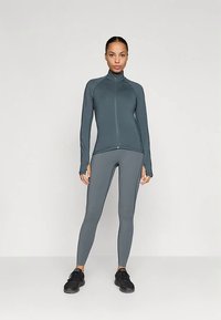 Mulher em pé ao centro, vestindo uma jaqueta cinza ajustada com fecho e leggings combinando, juntamente com ténis pretos, contra um fundo claro e simples.