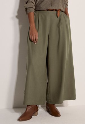 Donna con pantaloni olive a gamba larga con cintura marrone, maglione beige e stivaletti in pelle marrone, in piedi contro una parete chiara.