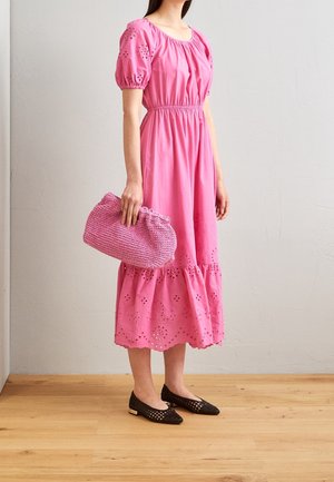 Kvinde i en lyserød midi-kjole med pufærmer og hulmønster, der holder en strikket lyserød clutch og bærer sorte vævede flade sko på trægulv.