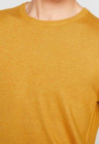 Senapsgul stickad tröja med rund halsringning och ribbade manschetter, som visar en slät textur och en subtil lyster.