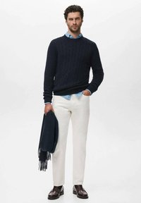 Suéter de punto trenzado en navy, camisa azul clara, pantalones crema y zapatos marrón oscuro. Sosteniendo una chal oscuro navy. Diseño limpio y moderno.