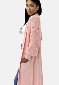 Lång, öppen frontkofta i ljusrosa, stickad struktur, med sluttande axlar och stora framfickor. Stylad över en vit topp och denim.