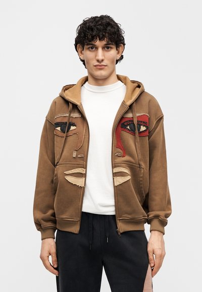 Καφέ φούτερ με φερμουάρ και κουκούλα, με κεντημένα σχέδια ματιών σε κόκκινο και μαύρο, και υφασμάτινες ταμπά λεπτομέρειες στο στήθος.