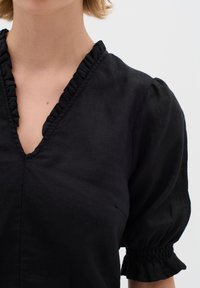 Schwarzes Leinenoberteil mit einem rüschigen V-Ausschnitt und puffärmeligen, gerafften Ärmelabschlüssen, das aus einem strukturierten, leichten Stoff besteht.