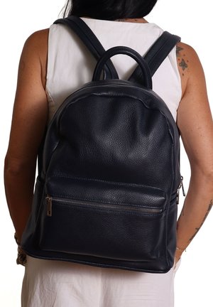 Femme portant un haut sans manches blanc, portant un sac à dos en cuir noir texturé avec une poche zippée à l'avant, avec des tatouages visibles sur le bras.