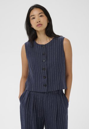 Femeie purtând un top bleumarin cu dungi subțiri, fără mâneci, cu nasturi, asortat cu pantaloni cu talie înaltă și dungi subțiri, cu mâinile în buzunare, pe un fundal neutru.