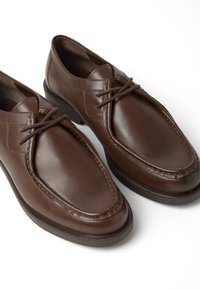 Des chaussures en cuir marron présentent un bout arrondi, un design à lacets et des détails de couture visibles le long des bords. Semelle en caoutchouc texturé.