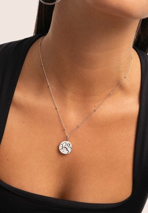 Collana d'argento con pendente circolare testurizzato che presenta un design di impronta di zampa. La catena ha piccole perline decorative, indossata su uno sfondo di tonalità della pelle.