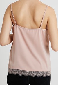Camisole rose clair avec des bretelles fines et ajustables, présentant une délicate dentelle au hem et un tissu lisse et texturé.