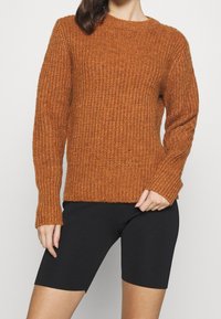 Brunt orange stickad tröja med ribbat mönster, rund halsringning och lång ärm. Matchad med svarta cykelbyxor, vilket ger en avslappnad stil.