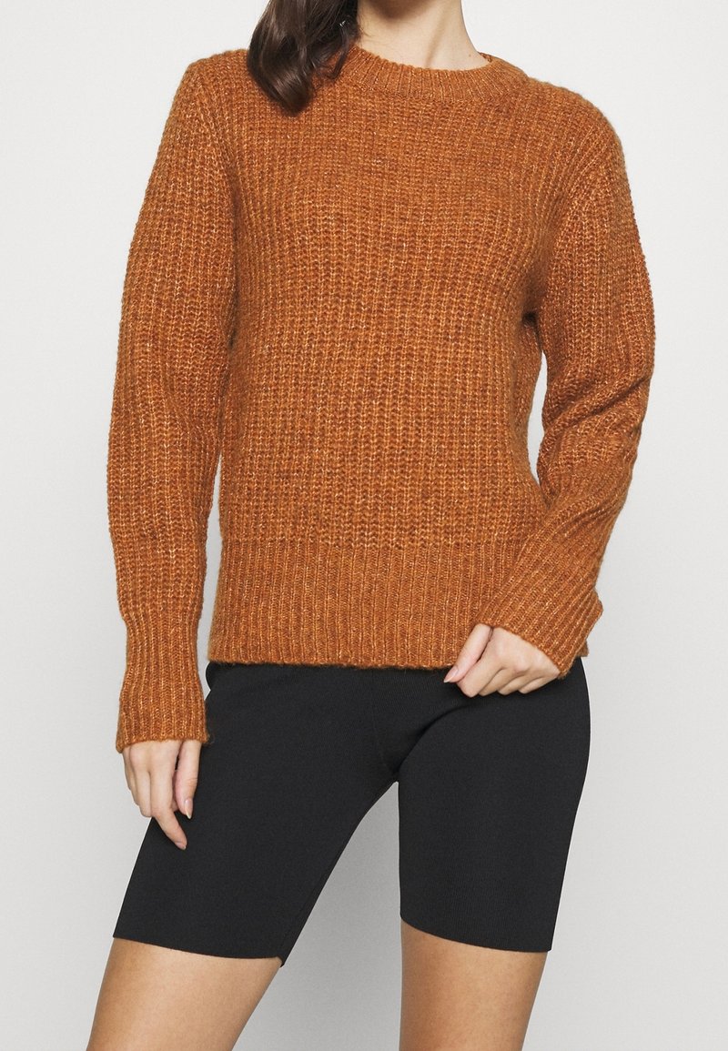 Brunt orange stickad tröja med ribbat mönster, rund halsringning och lång ärm. Matchad med svarta cykelbyxor, vilket ger en avslappnad stil.