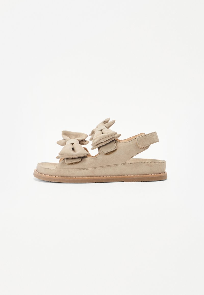 Beige suede sandals with two oversized bows on the front, open back design, flat sole, and stitched detailing along the edges.