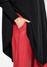 Pull en tricot noir à coupe ample avec fente sur le côté, associé à un pantalon en cuir rouge lisse avec une poche latérale.