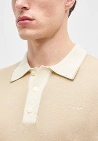 Gros plan sur une personne portant un polo en maille beige avec un col crème et trois boutons, affichant le logo GANT brodé sur la poitrine.