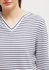 Donna che indossa un maglione a righe bianco e blu navy con scollo a V e cappuccio, mostrata dalla neck alla parte centrale del torso su uno sfondo semplice.