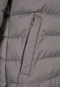 Grå jakke med puffer-design og quiltede sektioner, der har en lynlåslomme. Materialet har en glat tekstur med et let udseende.
