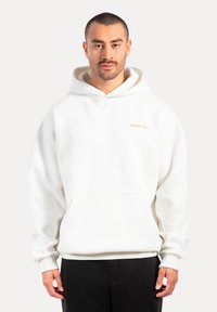 Uomo che indossa un hoodie bianco con tasca frontale e logo "Smilodox Sport", abbinato a pantaloni neri, in piedi davanti a uno sfondo semplice.