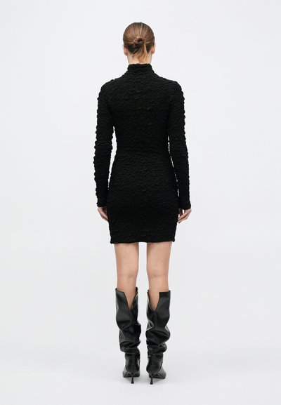 Naine, kellel on juuksed kuklas, kannab tekstuurse musta pika varrukaga lühikest kleiti ja musti lõdvalt istuvaid põlvikuti kõrge kontsaga saapaid, seisab seljaga vaataja poole.