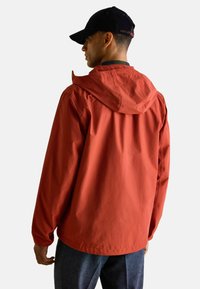 Vīrietis, tērpies rūsaini oranžā kapucē, tumšā cepurē un tumšās biksēs, stāvot ar muguru pret vienkāršu fonu.