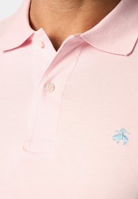 Světle růžová polo košile s klasickým límcem, obsahující jeden knoflík a malou modrou vyšívanou nášivku na levé části hrudníku.