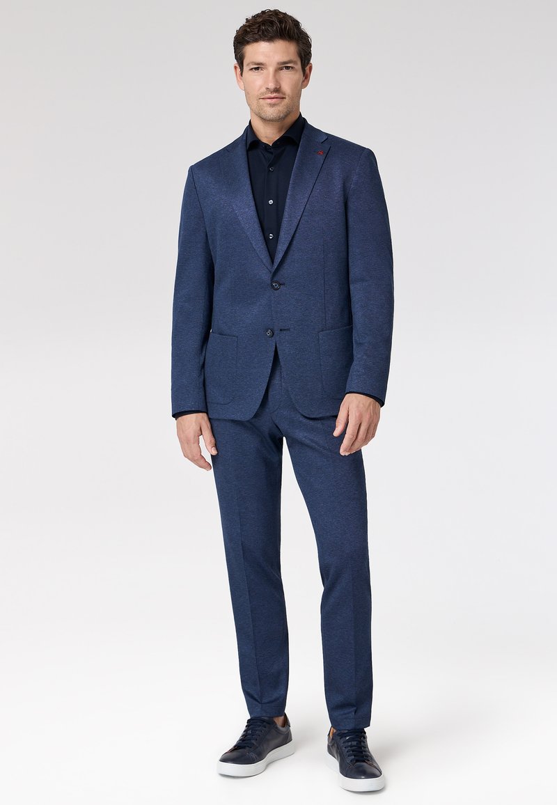 Marineblauw pak met een gestructureerde stof, een colbert met enkele rij knopen, met inkeping revers en twee voorzakken, gecombineerd met een donker overhemd en sneakers.