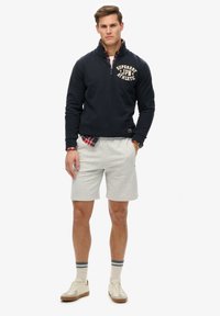 Felpa con cappuccio blu navy e mezza zip con stampa del logo, abbinata a pantaloni corti grigi chiari. Il modello indossa sneakers beige e calzini a righe. Camicia a quadri in vista.
