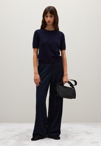 Marineblauwe korte mouwen trui, marineblauwe brede broek en een kleine zwarte handtas. Geverfde zwarte schoenen met pailletjes. Neutrale achtergrond.