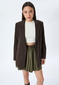 Bruine dubbele blazer met opstaande revers, gedragen over een witte cropped shirt, gecombineerd met een olijfgroene geplooide rok en een bruine riem.