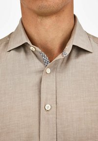 Camicia di lino beige con colletto appuntito, caratterizzata da un dettaglio interno del colletto a motivo. Due bottoni visibili sulla parte anteriore.