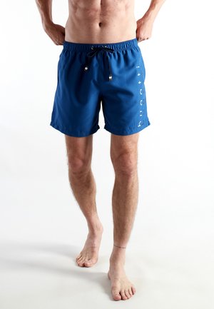 Man wearing blue swim shorts with drawstring, standing barefoot against a white background, torso and legs visible.